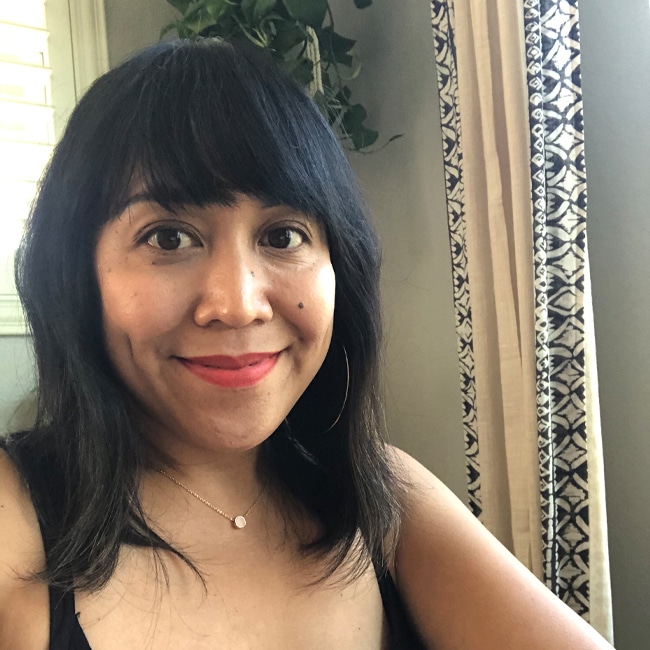 A person with long dark hair and hoop earrings is smiling at the camera. They are wearing a necklace and red lipstick, seated near a window with patterned curtains and a plant in the background.