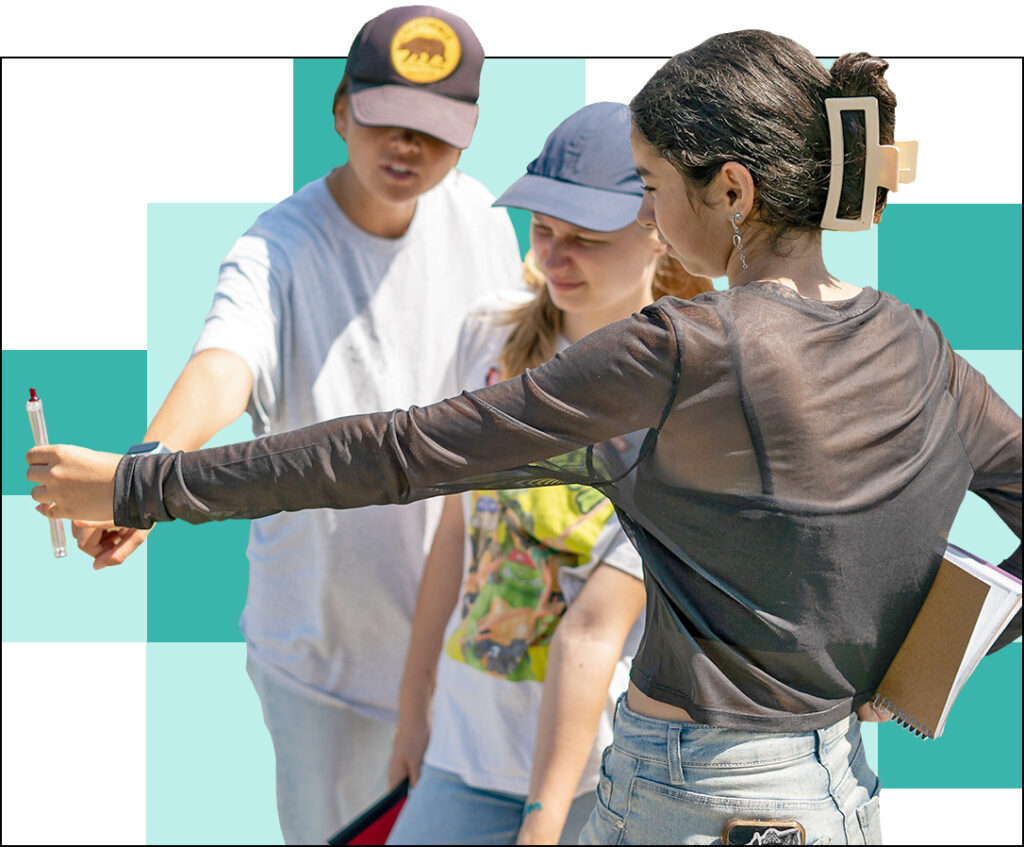 Three people outdoors; one is holding a pen and phone, possibly taking a selfie. The second person points at something, while the third smiles. They are casual and wearing hats, capturing a moment of professional development against a teal and white geometric pattern in the background.