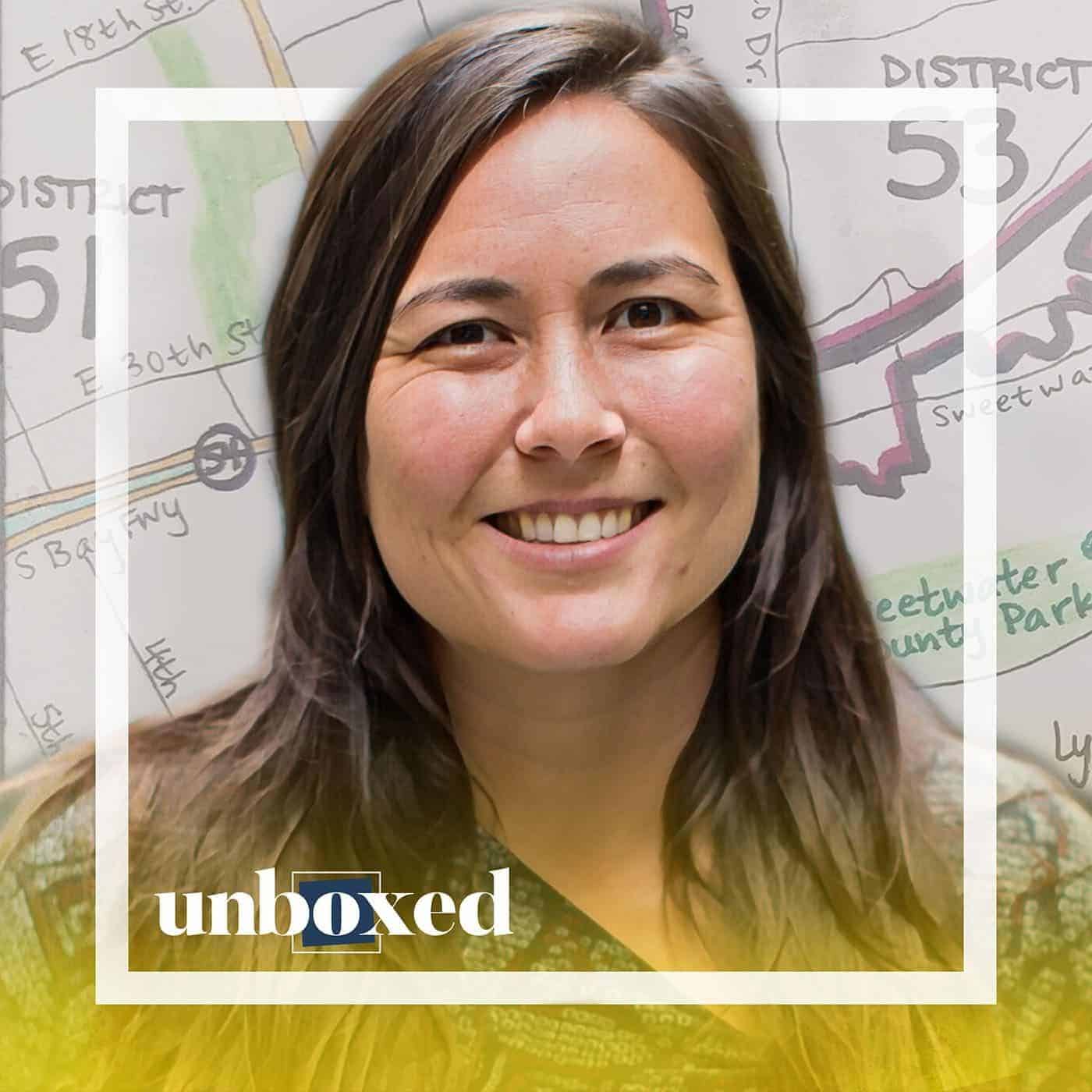A smiling woman with long brown hair is framed by a white border against a background featuring a map with district numbers, highlighting the nuances of redistricting. The word unboxed is overlaid at the bottom left of the image.