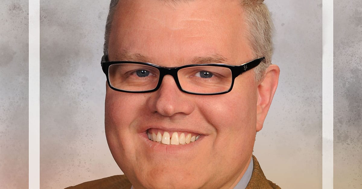 A smiling person with short hair and glasses exudes leadership while wearing a brown blazer. The backdrop is a neutral, textured gray, reminiscent of the deeper learning environment championed by visionaries like Scott McLeod.